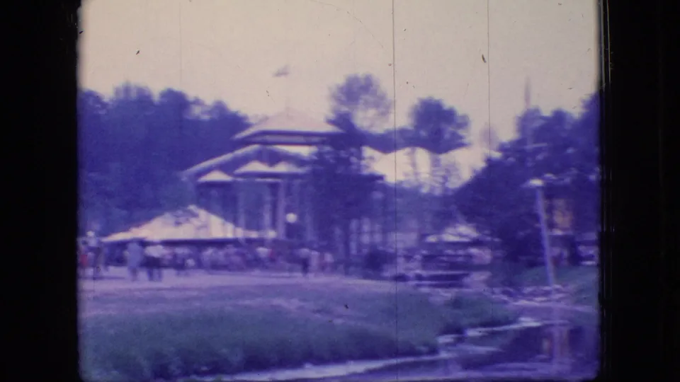 1966 Ohio home-movie frame showing a park structure, distant visitors, trees, lakeside grass, and a small dock or shoreline feature.