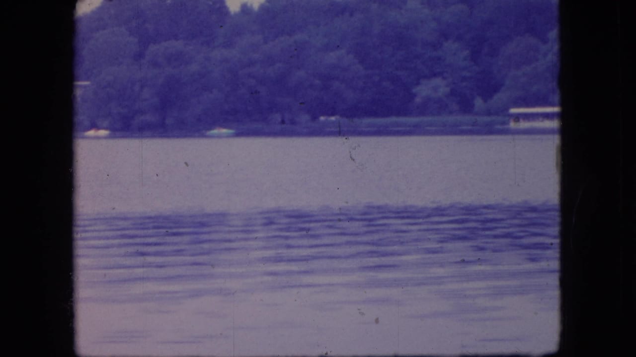 1966 Ohio 8mm frame of a quiet lake with rippled water, wooded shoreline, and small distant boats or pale objects near the far bank.