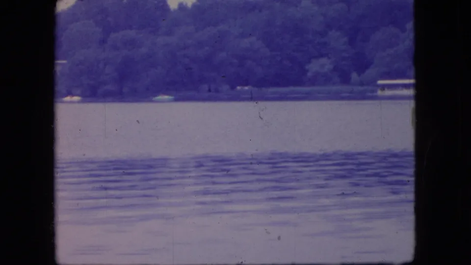 1966 Ohio 8mm frame of a quiet lake with rippled water, wooded shoreline, and small distant boats or pale objects near the far bank.