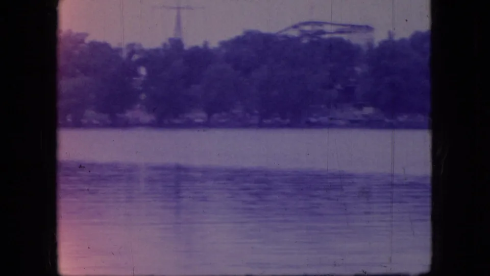 1966 Ohio 8mm frame of calm lake water and a wooded far shore, with a faint tower or vertical structure visible beyond the trees.
