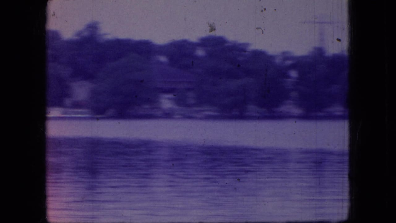 1966 Ohio 8mm frame showing a calm lake or broad water surface with a distant tree line, purple color shift, dust marks, and dark scan edges.