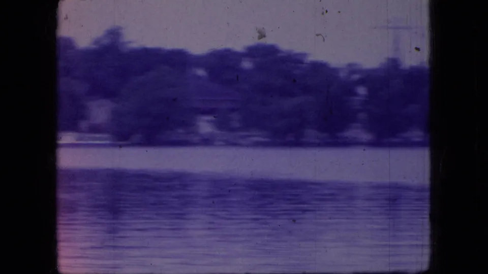 1966 Ohio 8mm frame showing a calm lake or broad water surface with a distant tree line, purple color shift, dust marks, and dark scan edges.