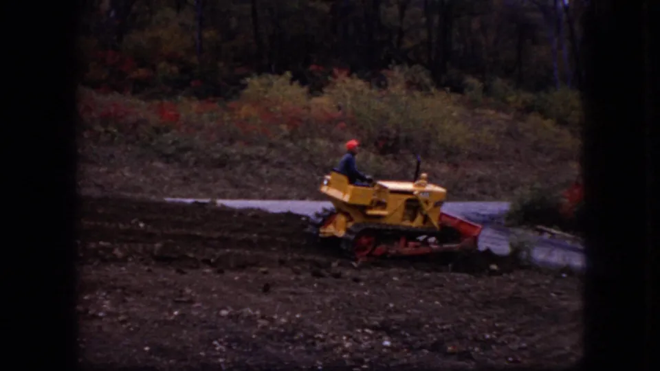 1966 Ohio 8mm frame showing a man in a red hard hat driving a yellow bulldozer across dark soil near a road and autumn brush.