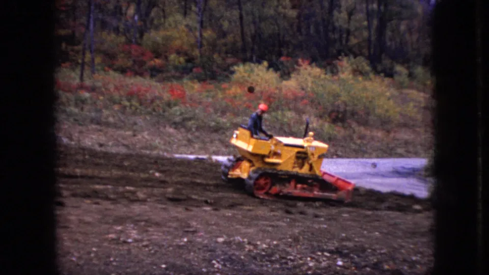 1966 Ohio 8mm frame of a hard-hatted operator on a yellow bulldozer beside a paved or light-colored path, with brush and trees in the background.