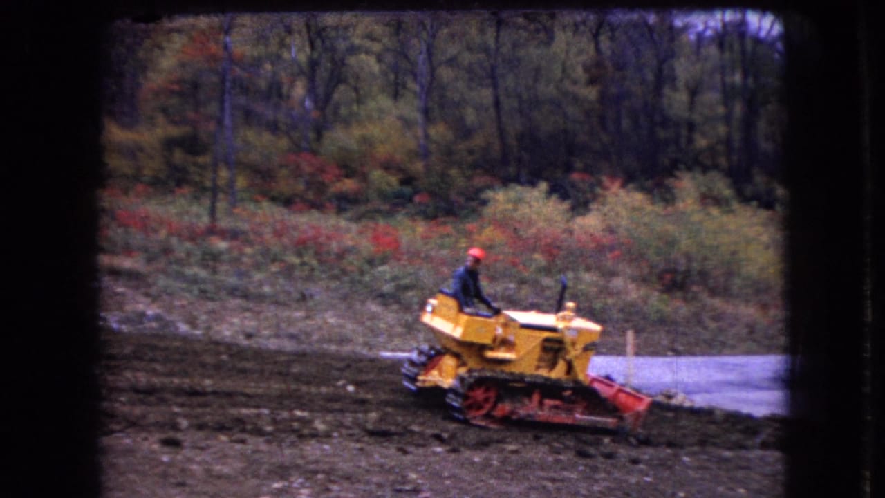 In 1966 Ohio home-movie footage, a man in a red hard hat operates a yellow tracked bulldozer on a muddy construction site bordered by autumn woods.