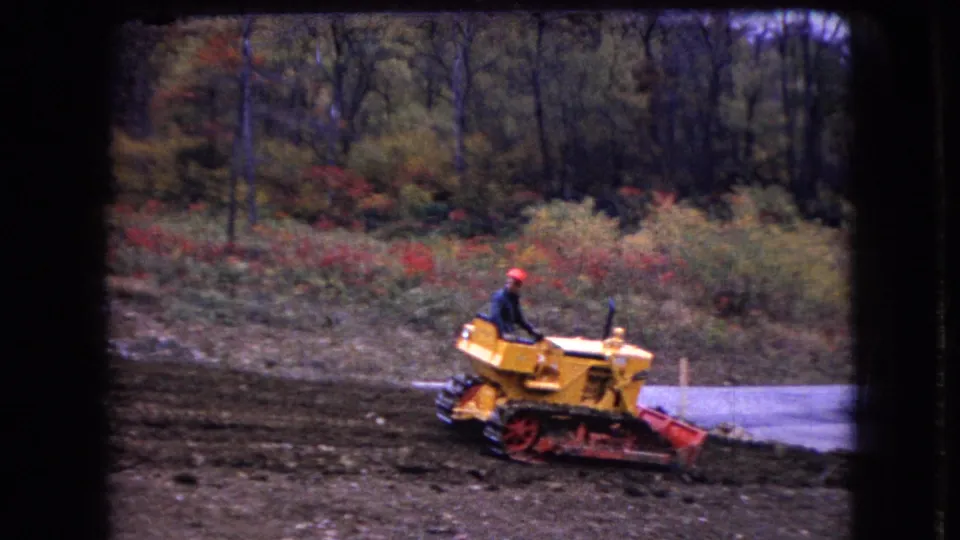 In 1966 Ohio home-movie footage, a man in a red hard hat operates a yellow tracked bulldozer on a muddy construction site bordered by autumn woods.