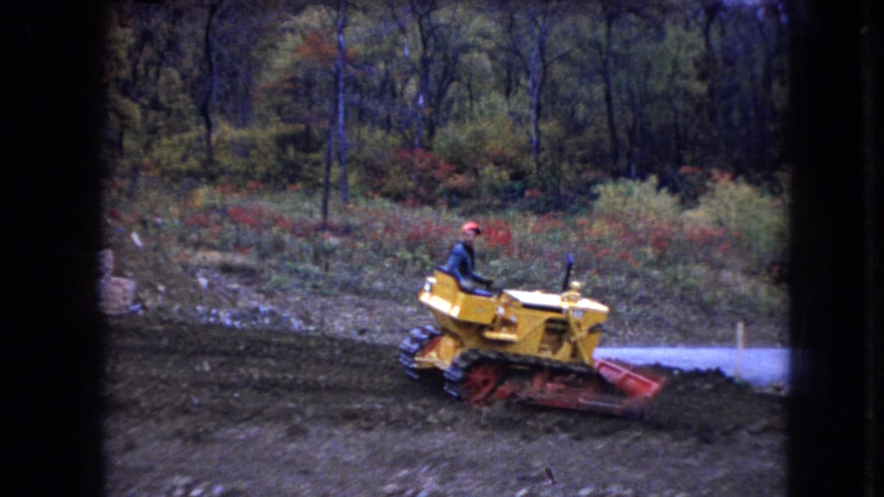 Yellow bulldozer with a seated operator moving beside a pale road and wooded edge.