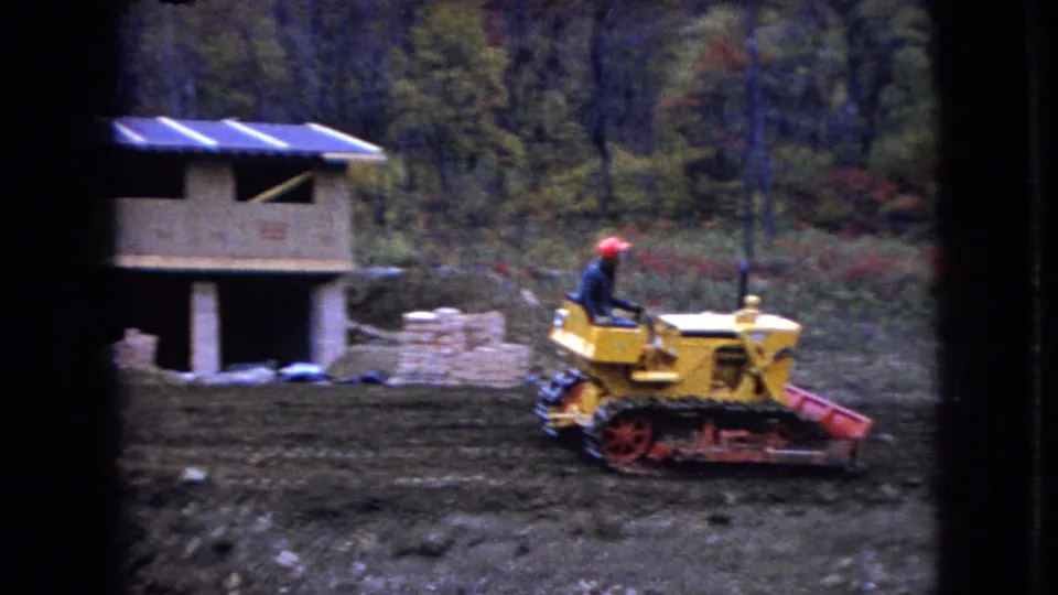 A man operates the yellow bulldozer near a partially built wooden structure and stacked materials in the 1966 Ohio construction-site footage.