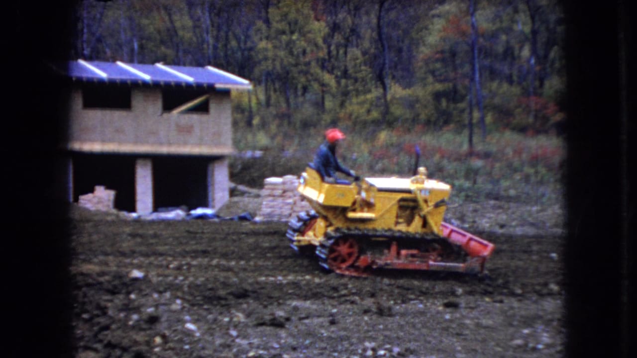 In 1966 Ohio home-movie footage, a man in a red hard hat operates a yellow tracked bulldozer beside an unfinished wooden building at a muddy rural construction site.