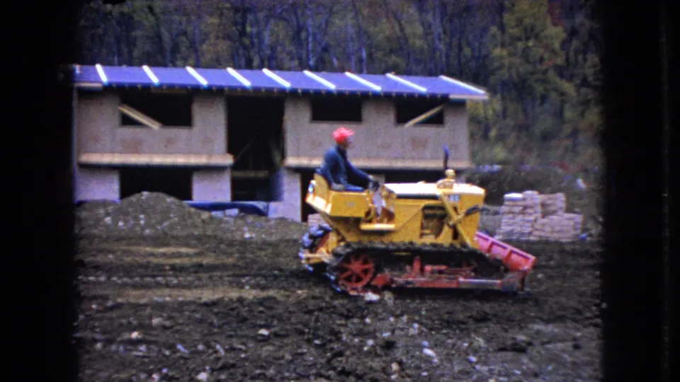 A man operates the yellow tracked bulldozer in front of an unfinished wooden structure during a 1966 Ohio rural construction scene recorded on 8mm film.