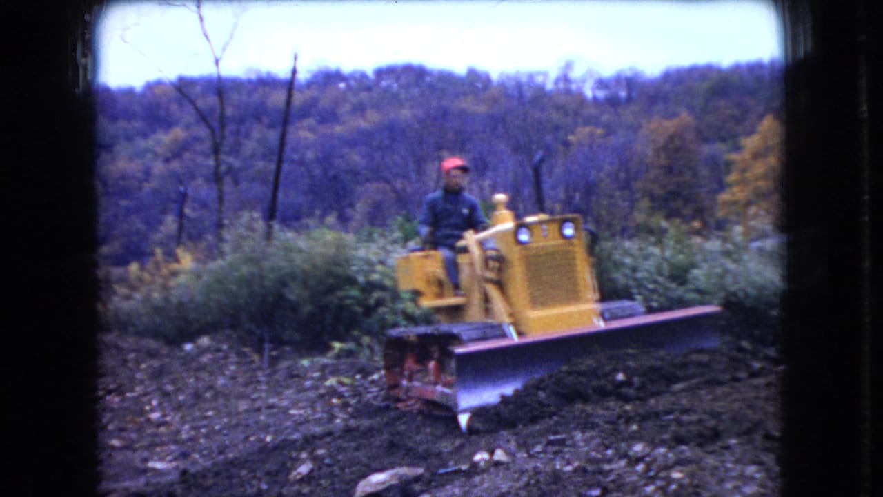 In 1966 Ohio home-movie footage, a man in a red cap drives a yellow Case bulldozer across uneven dirt with autumn trees behind him.