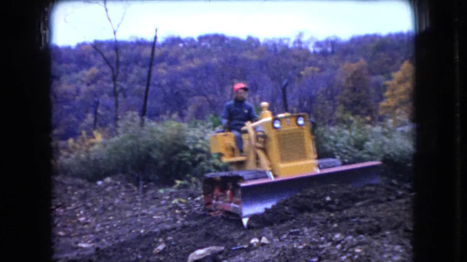 In 1966 Ohio home-movie footage, a man in a red cap drives a yellow Case bulldozer across uneven dirt with autumn trees behind him.