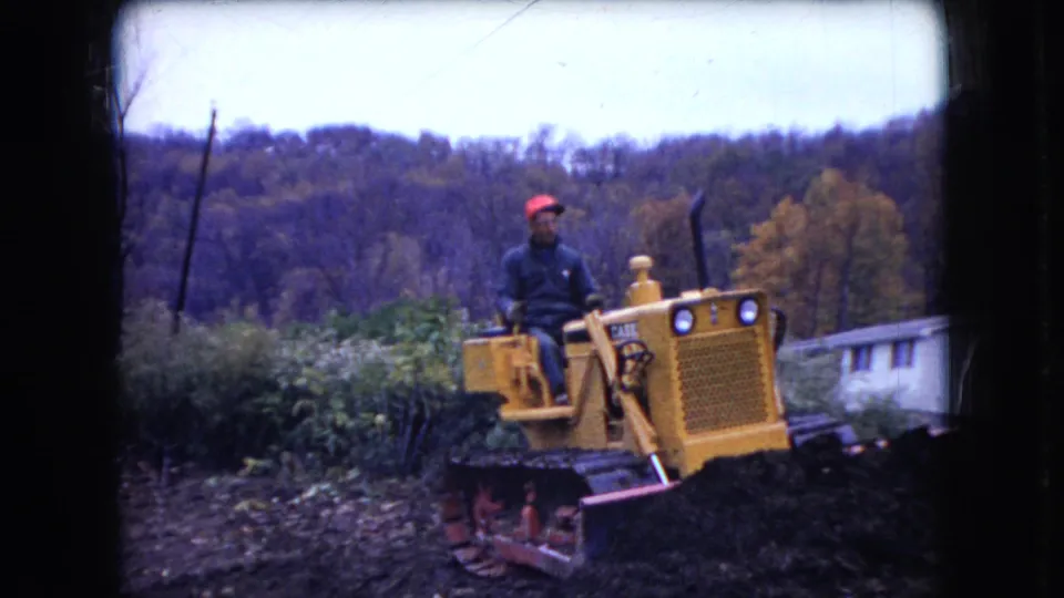 The operator sits on a yellow Case bulldozer with its blade low as it moves through rocky ground beside brush and a white house in Ohio, 1966.