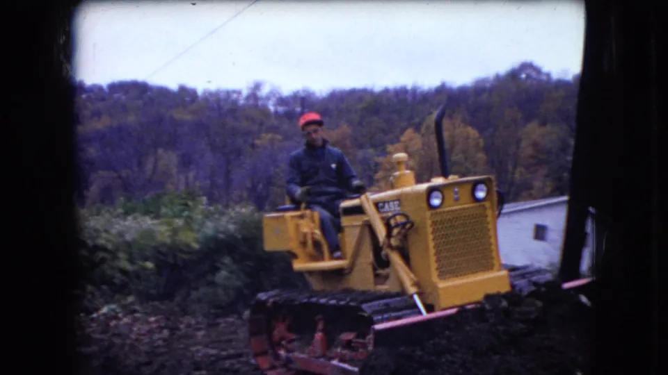 A man in a red cap steers the yellow bulldozer past churned soil, brush, and a white house, with a wooded autumn hillside in the background.