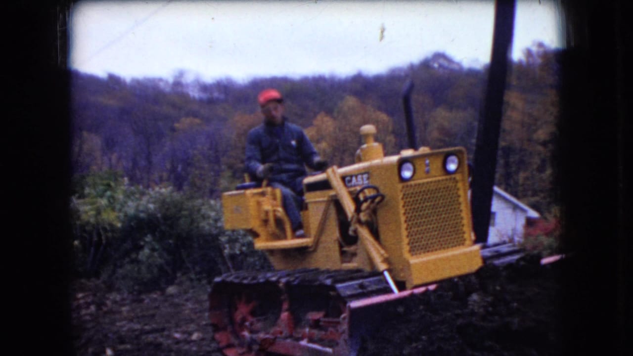 In this 1966 Ohio home-movie frame, a laborer in a red cap operates a yellow Case tracked bulldozer near autumn trees and churned ground.