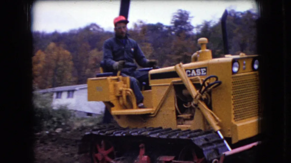 A laborer in dark clothing drives a yellow Case bulldozer, with autumn trees and a partly visible white building in the background.