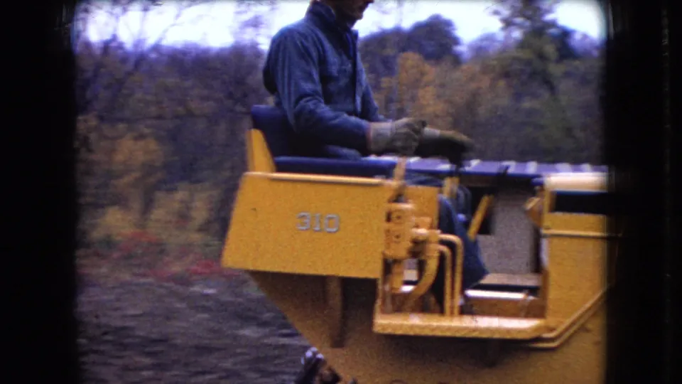 1966 Ohio 8mm frame with the bulldozer operator in profile, seated on the yellow Case 310 while the machine advances through a wooded dirt area.