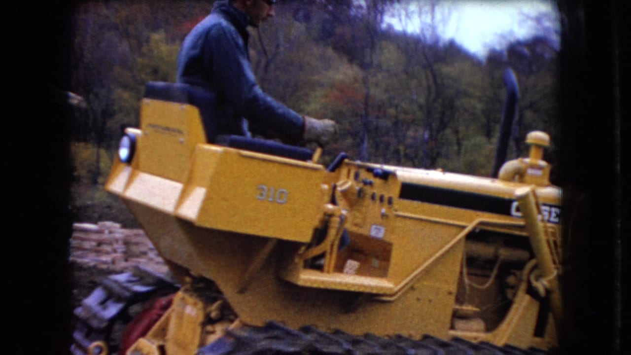 In 1966 Ohio home-movie footage, a man in a blue shirt operates a yellow Caterpillar 310 tracked bulldozer in a wooded area.