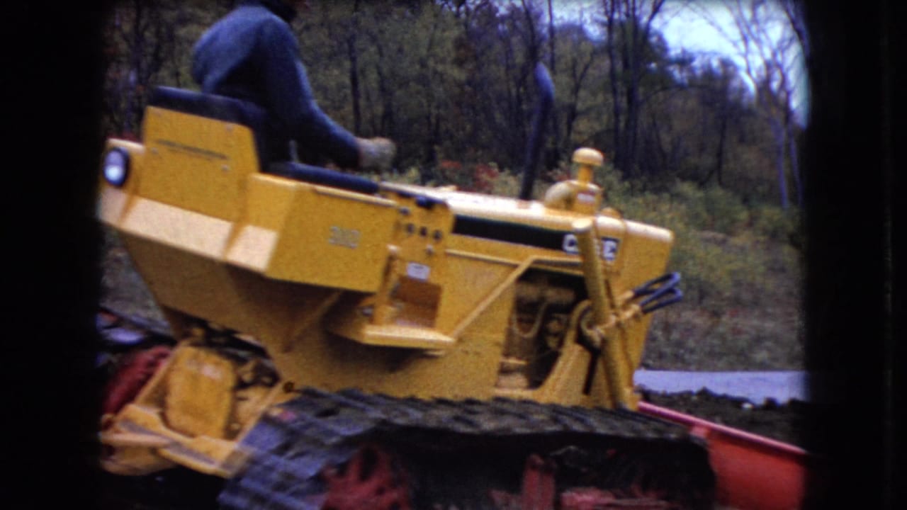 The yellow Caterpillar 310 bulldozer is seen from the side with its operator partly visible against an Ohio wooded backdrop.