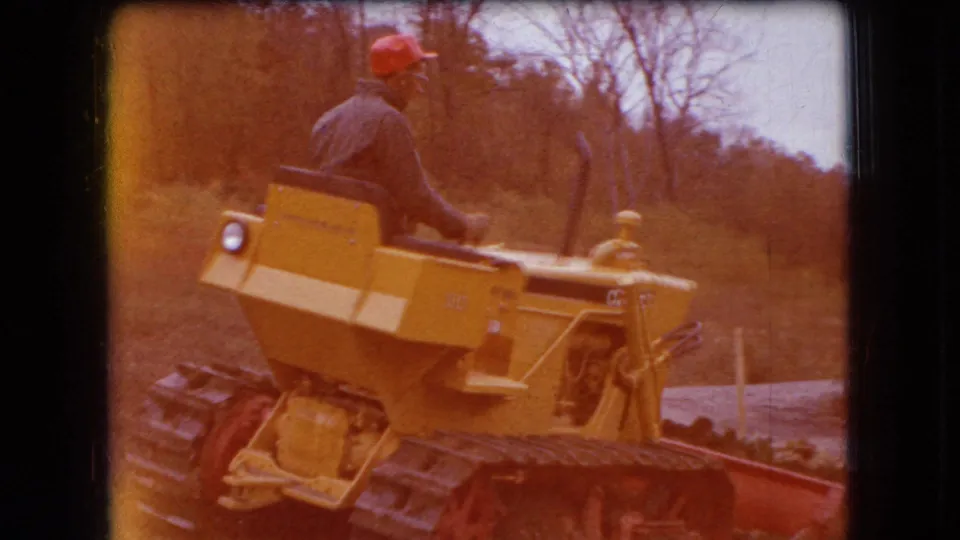 A man wearing a red cap and blue shirt drives the yellow tracked Caterpillar bulldozer through a wooded Ohio landscape in faded 8mm color.