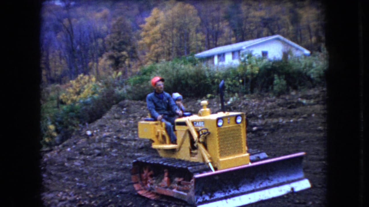 In supplied 1966 Ohio home-movie footage, a man in a red cap and another seated person ride a yellow tracked bulldozer across a rough rural yard below a white house and wooded hillside.