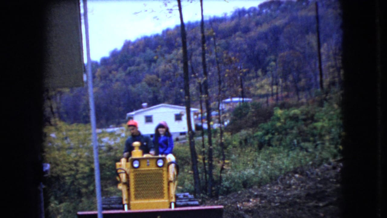 Two adults, one wearing a red cap and one in a blue jacket, ride a yellow bulldozer toward the camera in a rural Ohio yard with a white dwelling, utility pole, and wooded hillside behind them.