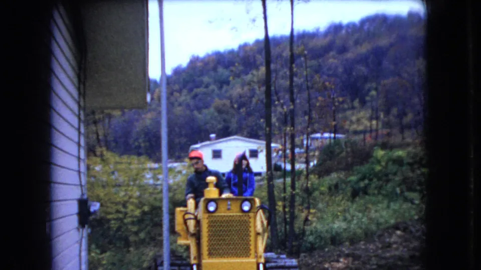The yellow bulldozer carrying two adults passes near the side of a house, with a utility pole, white dwelling, brush, and wooded hills visible in the 1966 Ohio home-movie frame.
