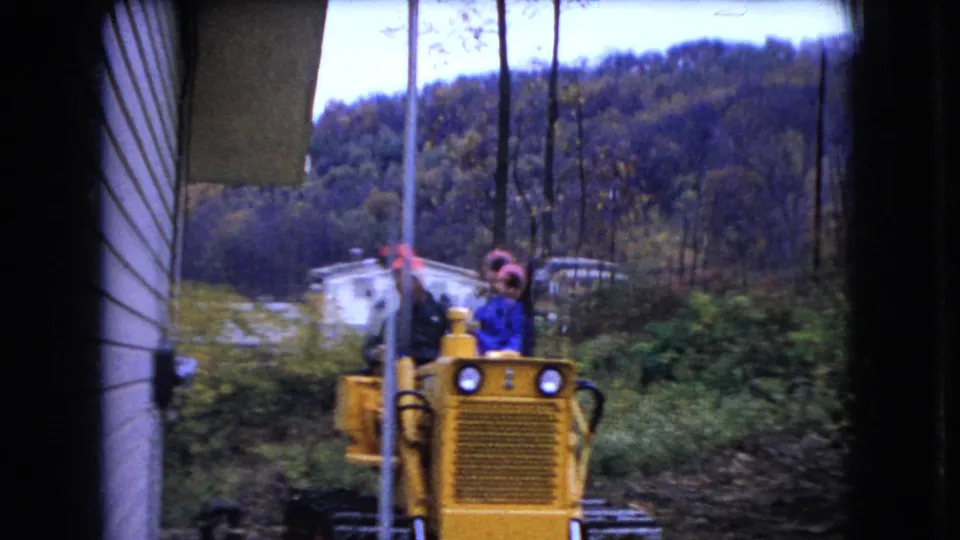 At the end of the short clip, two adults ride the yellow bulldozer near a utility pole, with a white dwelling, muddy track, brush, and autumn hillside behind them in rural Ohio.