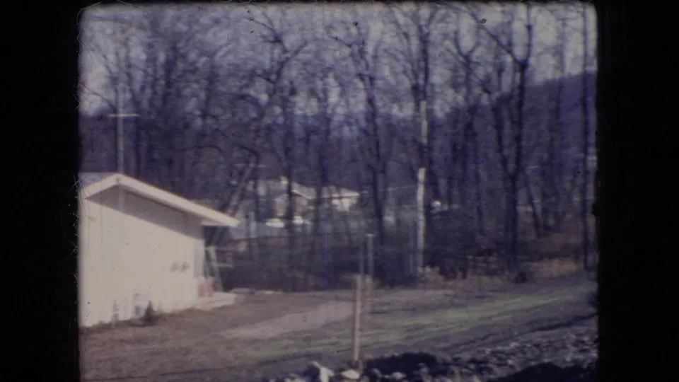 Passing 1966 Ohio roadside view of the side of a light-colored house, a driveway, lawn, utility poles, and bare trees on a hillside.