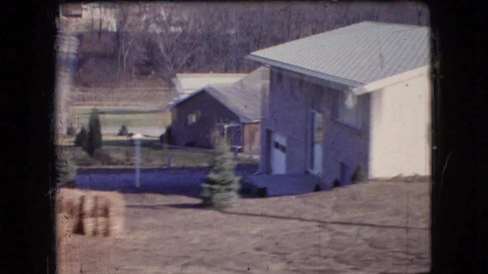 1966 Ohio farm-country view showing the rear or side of a light-colored house, a darker outbuilding, small evergreens, hay bales, lawn, and leafless trees.