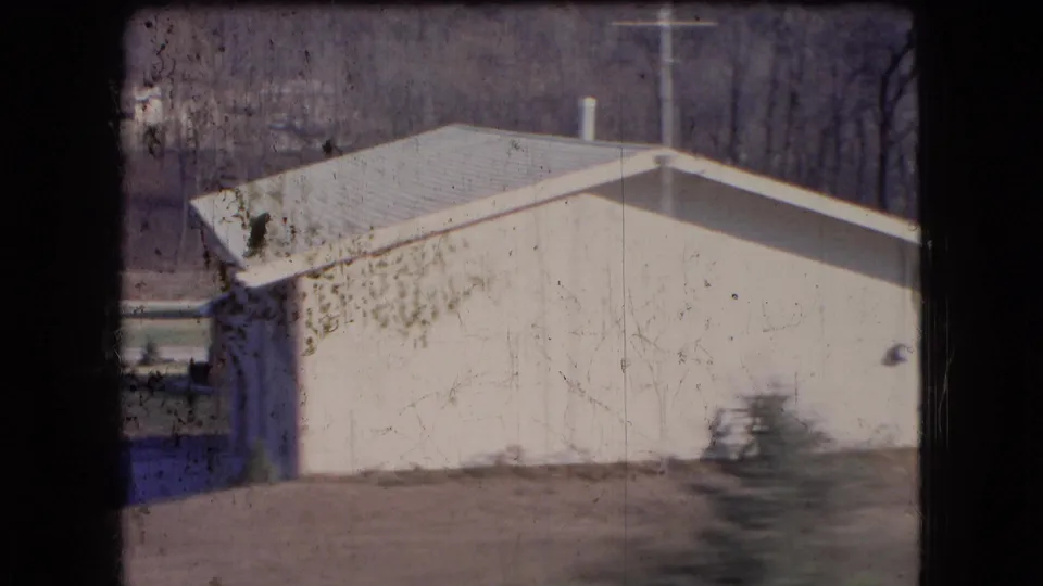 Close roadside frame from 1966 Ohio showing a light-colored single-story house with a metal roof, bare trees, utility pole, small evergreen, and film edge marks.