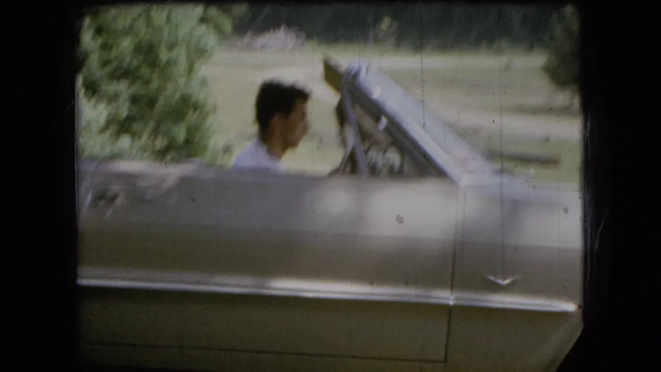 In 1966 Ohio home-movie footage, a young man in a light shirt sits in a silver convertible with the top down beside grass and trees.