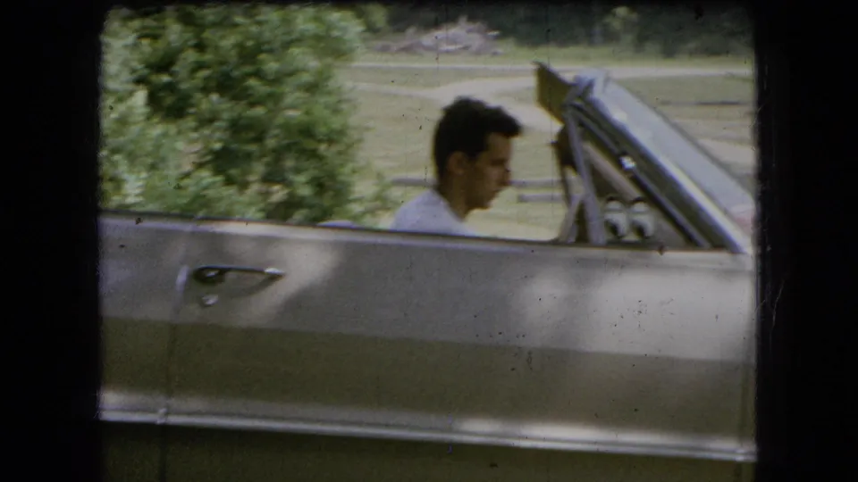 A side view shows the young driver in a light shirt seated low behind the door of a silver convertible in a green outdoor setting.