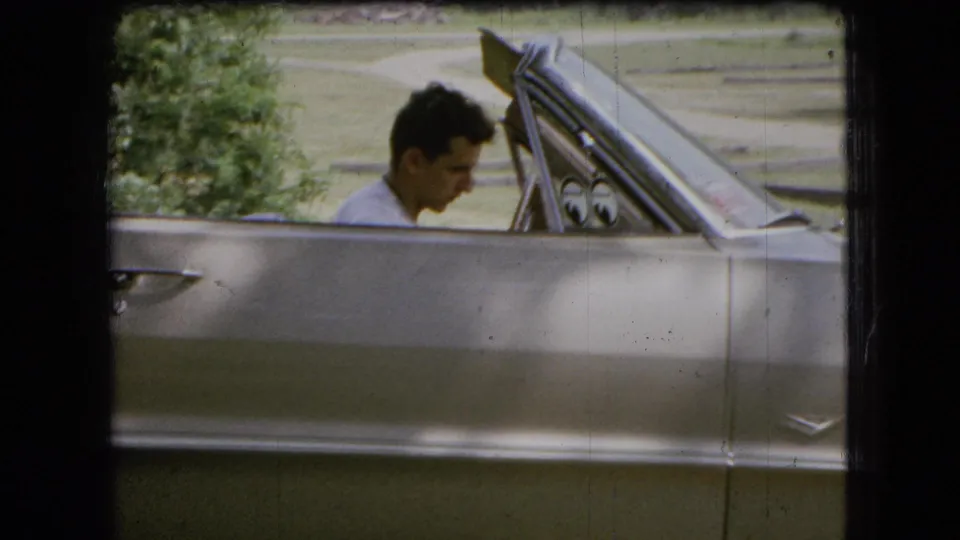 The driver appears to look down toward the controls or dashboard area while sitting in the silver convertible with greenery behind him.