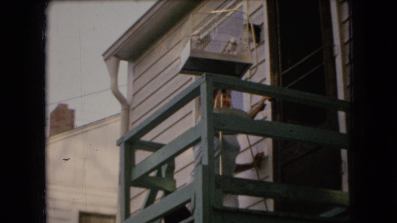 1966 Ohio home-movie frame showing an adult woman near a green back porch railing beside white house siding and an open doorway.
