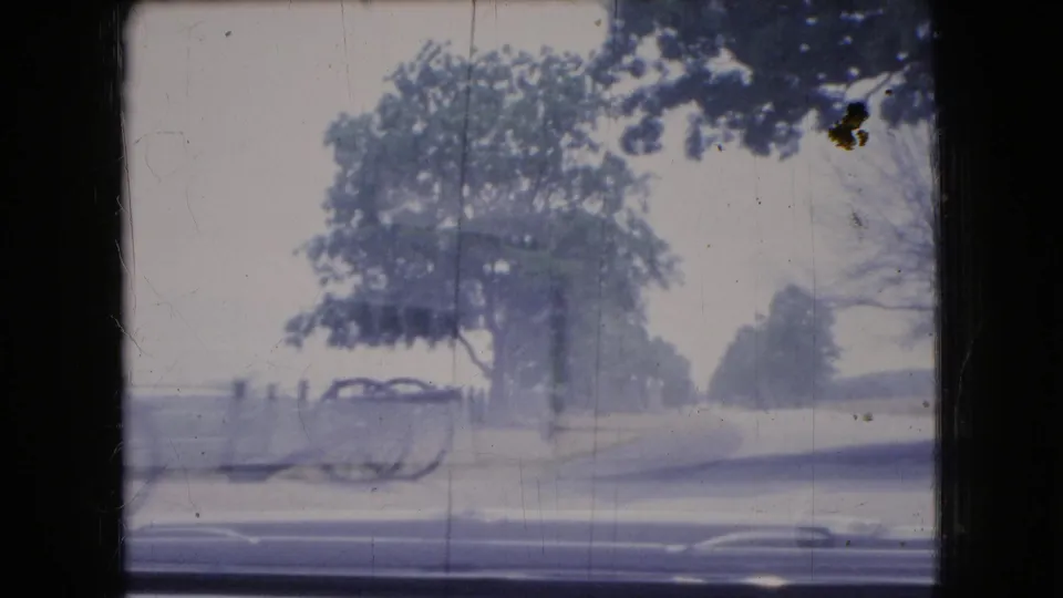 1966 Ohio home-movie frame with a moving-vehicle view of roadside trees, open pavement, and a classic car partly visible beyond the road.