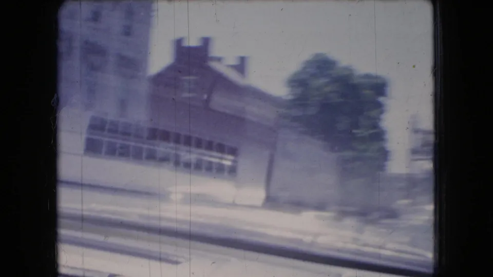 1966 Ohio 8mm frame showing a blurred street-side building facade, tree, pavement, and roadside curb from inside a moving car.