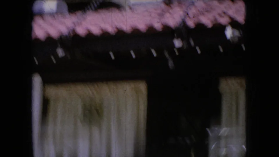1966 Ohio home-movie frame showing a dark roadside building facade with a red shingled roof and pale curtains, partially blurred by camera movement.
