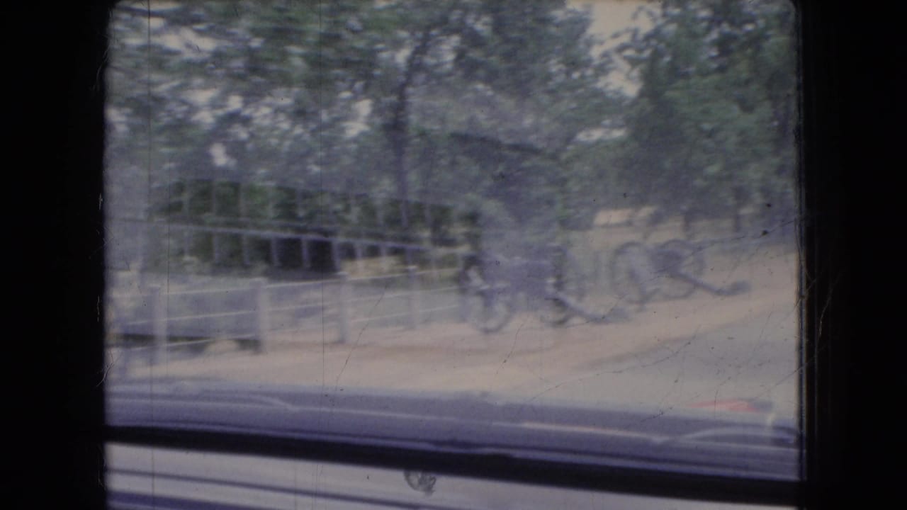 A 1966 Ohio home-movie frame shows blurred cannons and a park road viewed through a vehicle window, with trees and window framing visible.