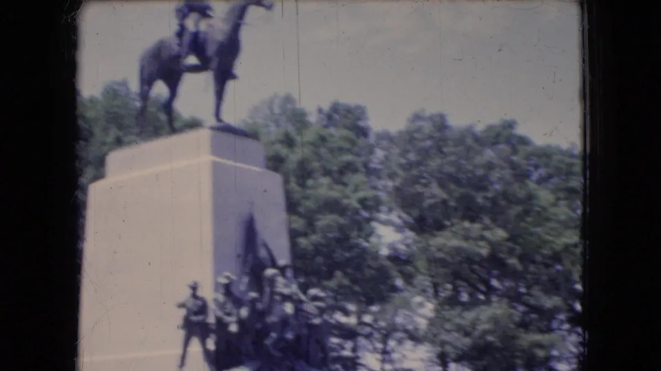 The 8mm frame shows the monument from a moving visitor viewpoint, with the horse-and-rider statue partly cropped above a stone pedestal and figures below.