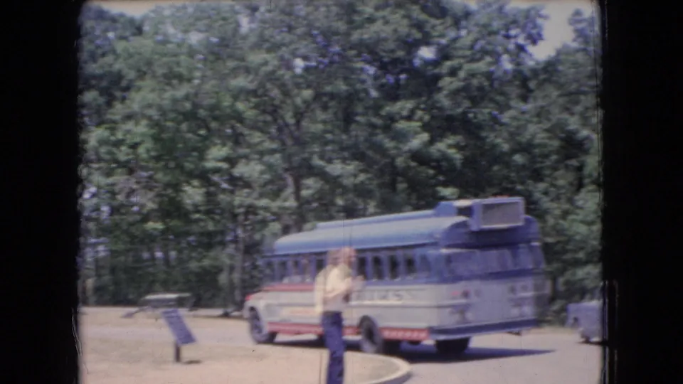 A blue mid-century tour bus is parked in a wooded Ohio site, with an adult visitor standing nearby and a sign or marker visible beside the road.