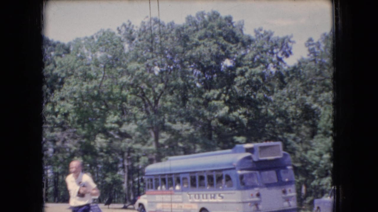 Man with camera passes in front of a parked tour bus beside dense trees.