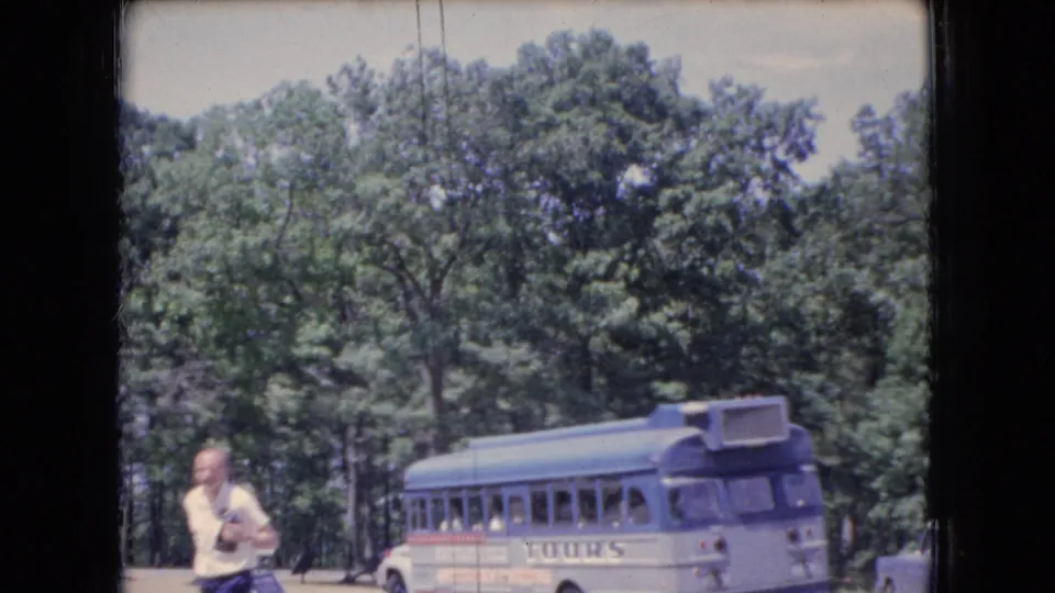 1966 Ohio home-movie frame with a casually dressed man and camera in front of a parked tour bus, backed by dense green trees and a bright sky.