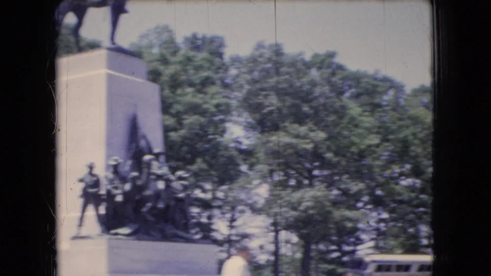 1966 Ohio home-movie frame showing part of a monument or sculptural base near trees, with the edge of the tour bus visible in the lower right.