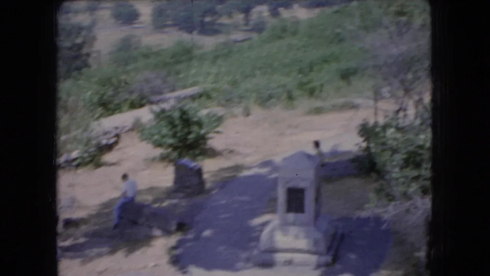 1966 Ohio cemetery view with a small stone monument or grave marker near a dirt path, trees, and a distant person in casual clothing.