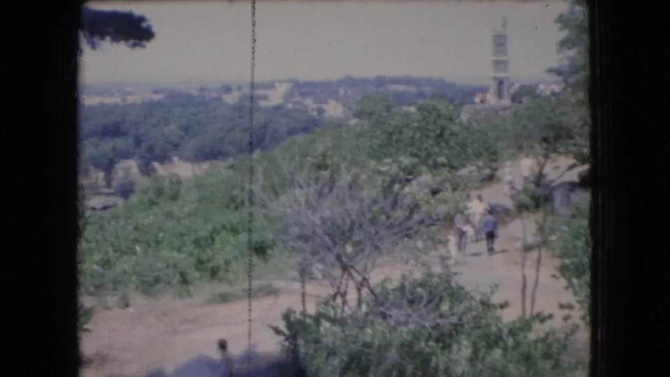 1966 Ohio home-movie frame looking across trees and a cemetery landscape toward distant visitors and an unidentified tower-like structure on the horizon.