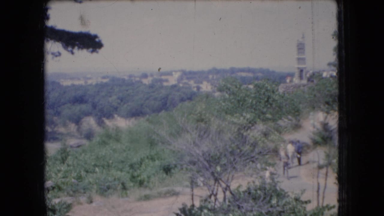 1966 Ohio 8mm frame showing a wooded valley overlook with a dirt path, a distant tower, and small visitors moving through the landscape.