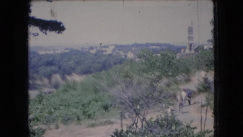 1966 Ohio 8mm frame showing a wooded valley overlook with a dirt path, a distant tower, and small visitors moving through the landscape.