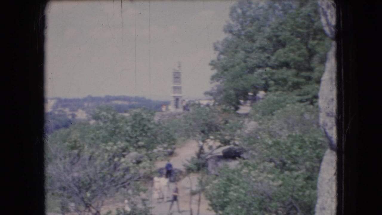 1966 Ohio home-movie frame of a tree-covered overlook with a winding dirt path, distant tower, and visitors partially obscured by foliage.