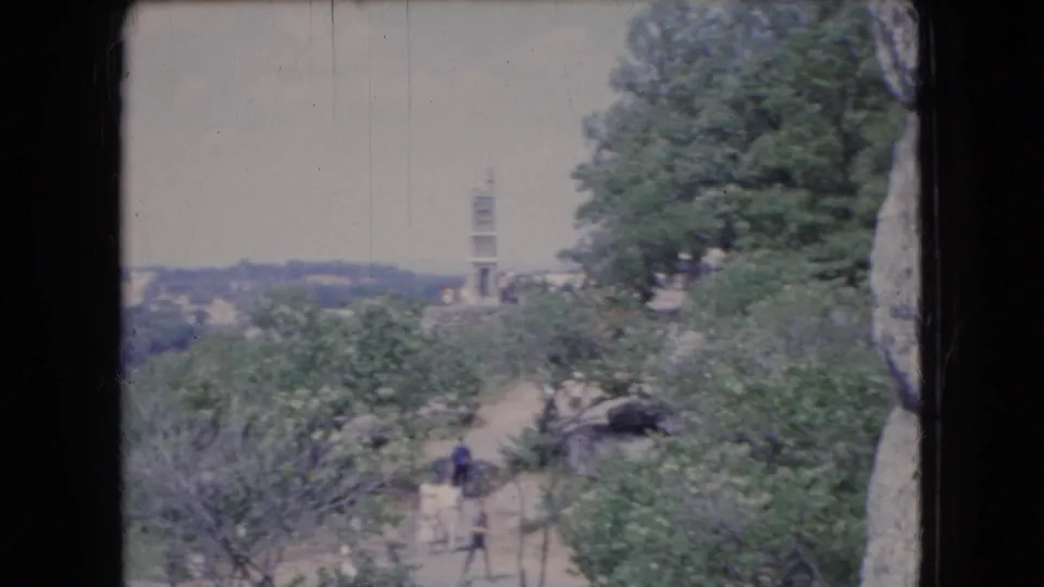 1966 Ohio home-movie frame of a tree-covered overlook with a winding dirt path, distant tower, and visitors partially obscured by foliage.